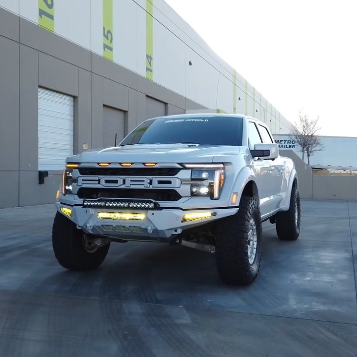 White Ford Raptor pickup with an aftermarket front bumper and LED light bar parked outside a row of shop units